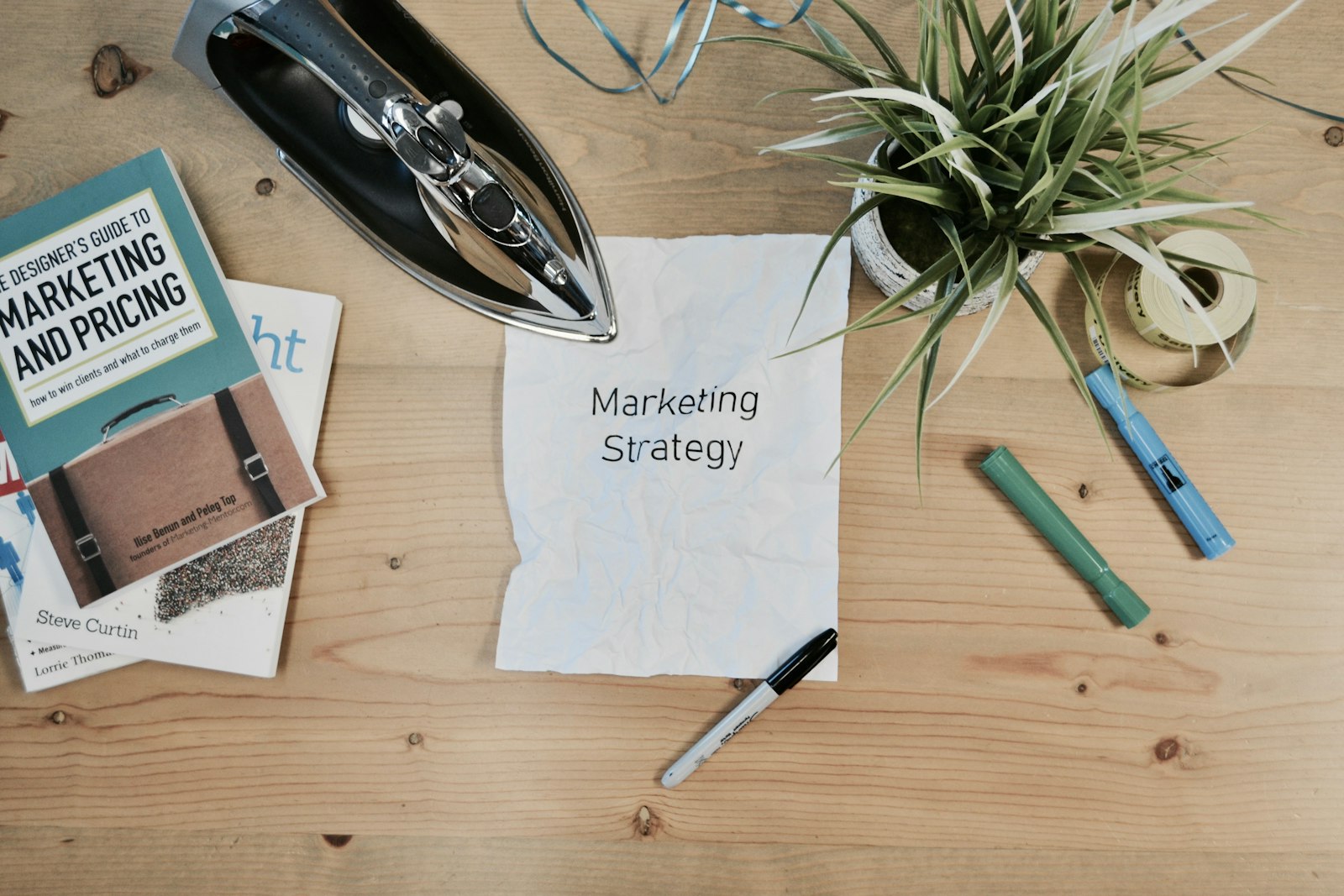 A wooden table with various items arranged on it including a crumpled piece of paper with the words 'Marketing Strategy', an iron, a pot plant, three markers, and two books. One of the books is titled 'A Designer's Guide to Marketing and Pricing'.