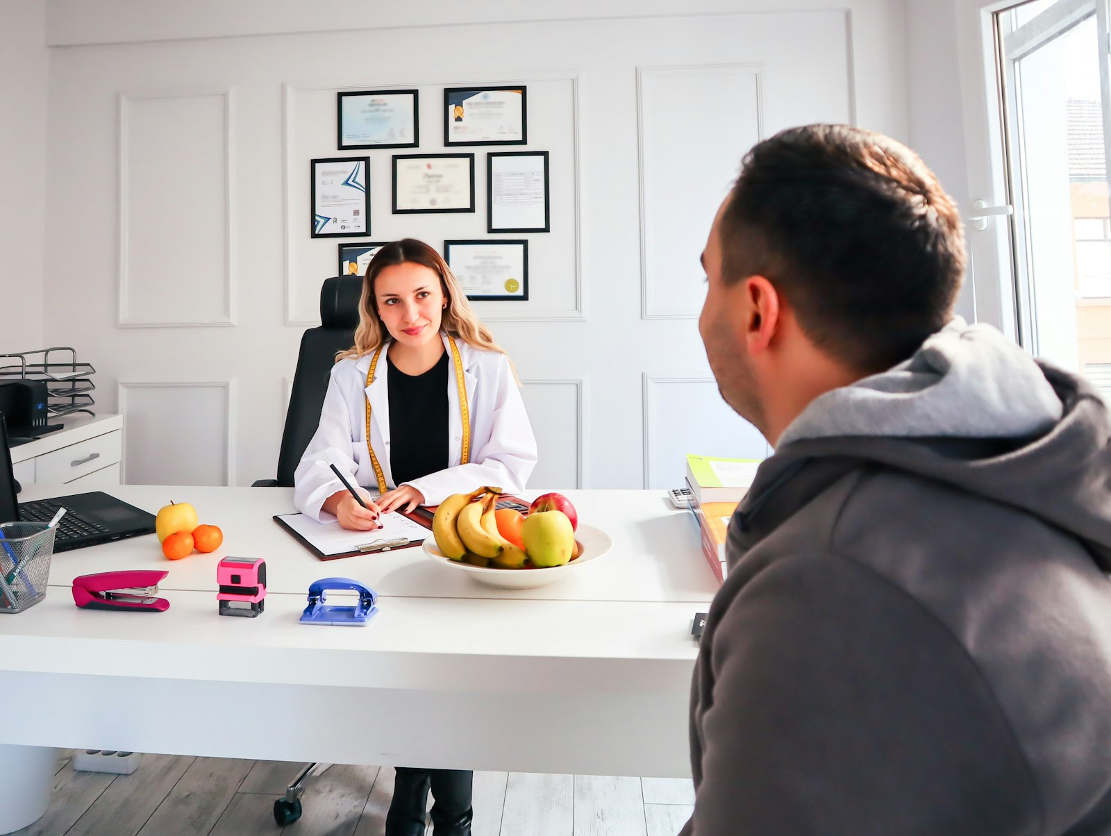 A professional consultation setting with a medical professional sitting at a desk facing a client. The room has a modern aesthetic with white walls decorated with framed certificates. The desk is organized with office supplies, a laptop, and a fruit bowl in the center.