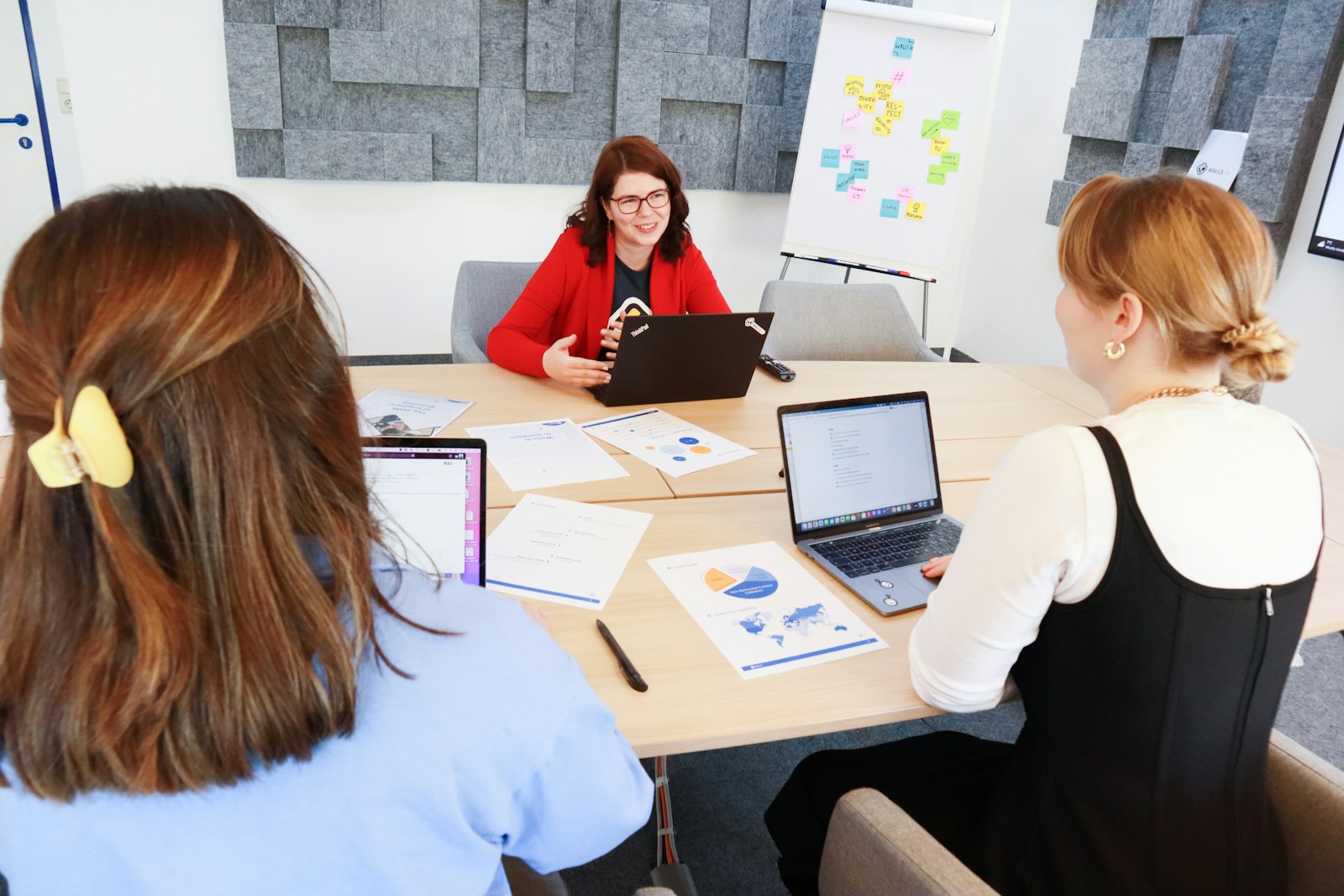 Three people are sitting around a conference table in a modern office. One person is wearing a red blazer and is speaking, while the other two listen and have open laptops in front of them. Papers with charts and graphs are scattered on the table. In the background, there are gray acoustic panels on the wall and a flip chart with colorful notes.